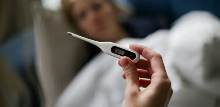 Hand holding a digital thermometer with blurred background of a sick person.