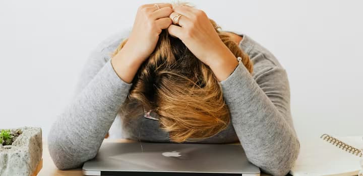 A woman in gray long sleeves resting her head on a laptop, appearing tired or stressed